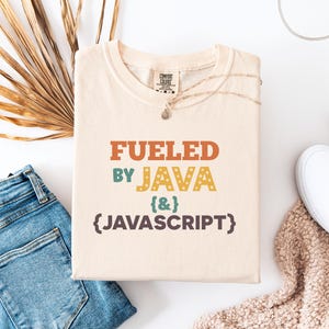 Op de afbeelding: Cr&egrave;mekleurig T-shirt met de tekst "Fueled by Java {&} Javascript" in oranje, blauwgroen, geel en bruin. Het shirt is opgevouwen en op een wit oppervlak geplaatst, naast een spijkerbroek, een ketting en witte sneakers.