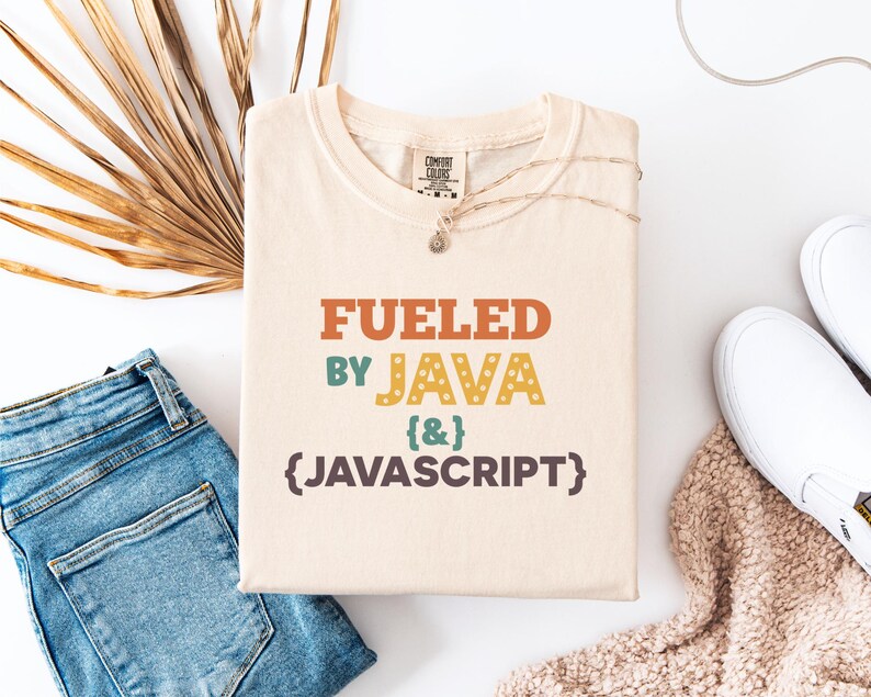 Op de afbeelding: Cr&egrave;mekleurig T-shirt met de tekst "Fueled by Java {&} Javascript" in oranje, blauwgroen, geel en bruin. Het shirt is opgevouwen en op een wit oppervlak geplaatst, naast een spijkerbroek, een ketting en witte sneakers.