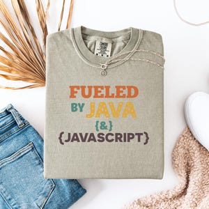 Op de afbeelding: Een saliegroen t-shirt met de tekst "Fueled by Java & Javascript" in oranje, geel, blauwgroen en bruin. Het shirt is opgevouwen en geaccessoireerd met een zilveren ketting, een blauwe spijkerbroek, witte sneakers en een beige deken.