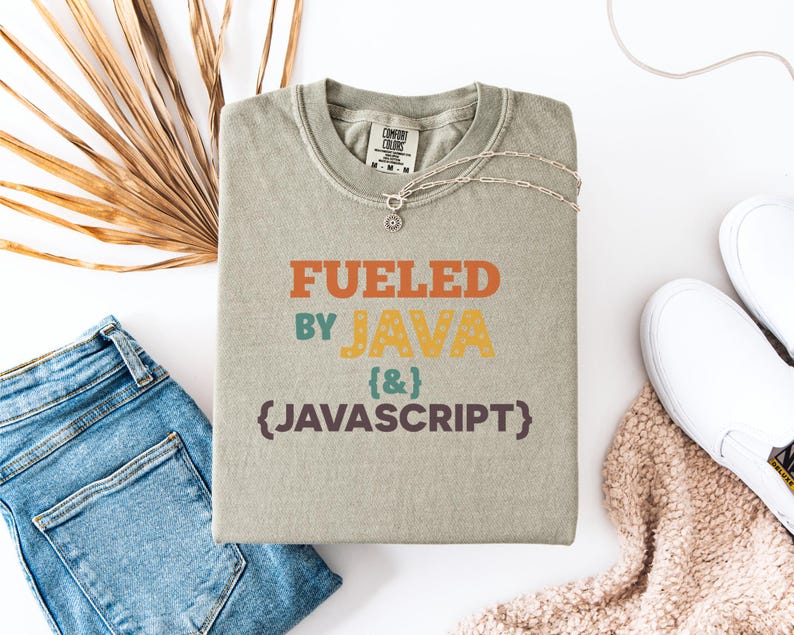 Op de afbeelding: Een saliegroen t-shirt met de tekst "Fueled by Java & Javascript" in oranje, geel, blauwgroen en bruin. Het shirt is opgevouwen en geaccessoireerd met een zilveren ketting, een blauwe spijkerbroek, witte sneakers en een beige deken.