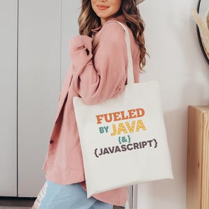 K&ouml;nnte beinhalten: Beige Tragetasche mit dem Text "Fueled by Java {&} Javascript" in Orange, Gr&uuml;n und Blau. Die Tasche wird von einer Person getragen, die eine rosa Jacke und blaue Jeans tr&auml;gt.