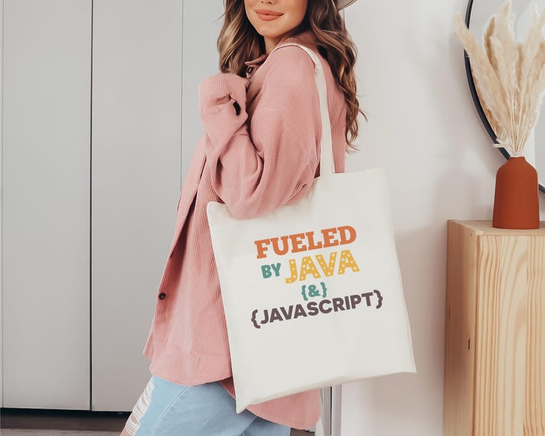 K&ouml;nnte beinhalten: Beige Tragetasche mit dem Text "Fueled by Java {&} Javascript" in Orange, Gr&uuml;n und Blau. Die Tasche wird von einer Person getragen, die eine rosa Jacke und blaue Jeans tr&auml;gt.