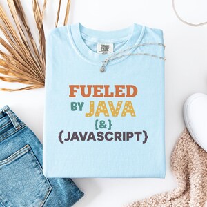 Op de afbeelding: Lichtblauw T-shirt met de tekst "FUELED BY JAVA & JAVASCRIPT" in oranje, groen en geel. Het shirt wordt gecombineerd met een blauwe jeans, witte sneakers en een zilveren ketting. Een casual, tech-thema ontwerp.