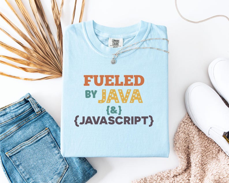 Op de afbeelding: Lichtblauw T-shirt met de tekst "FUELED BY JAVA & JAVASCRIPT" in oranje, groen en geel. Het shirt wordt gecombineerd met een blauwe jeans, witte sneakers en een zilveren ketting. Een casual, tech-thema ontwerp.