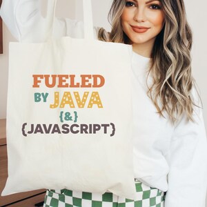 K&ouml;nnte beinhalten: Cremefarbene Tragetasche mit dem Text "FUELED BY JAVA {&} (JAVASCRIPT)" in Orange, T&uuml;rkis und Gelb. Die Tasche wird von einer Person gehalten, die einen wei&szlig;en Pullover tr&auml;gt. Der Hintergrund umfasst einen Holzschrank und ein gr&uuml;n-wei&szlig;es Karomuster.
