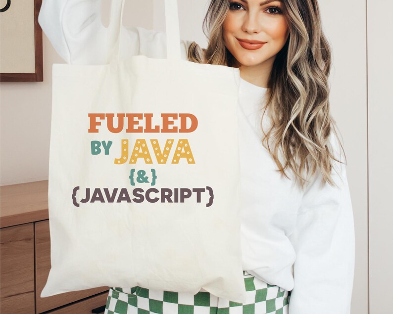 K&ouml;nnte beinhalten: Cremefarbene Tragetasche mit dem Text "FUELED BY JAVA {&} (JAVASCRIPT)" in Orange, T&uuml;rkis und Gelb. Die Tasche wird von einer Person gehalten, die einen wei&szlig;en Pullover tr&auml;gt. Der Hintergrund umfasst einen Holzschrank und ein gr&uuml;n-wei&szlig;es Karomuster.