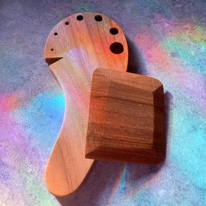 May include: Two wooden art tools, a palette and a square block, are displayed against a colorful background. The palette has several holes and a curved handle. The square block is a darker shade of brown.