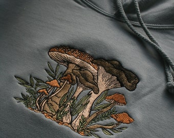 May include: A sage green hoodie with an embroidered design of mushrooms and foliage. The mushrooms are brown and white with orange accents.