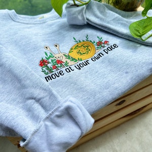 May include: A light gray sweatshirt with an embroidered snail design and the text "move at your own pace".