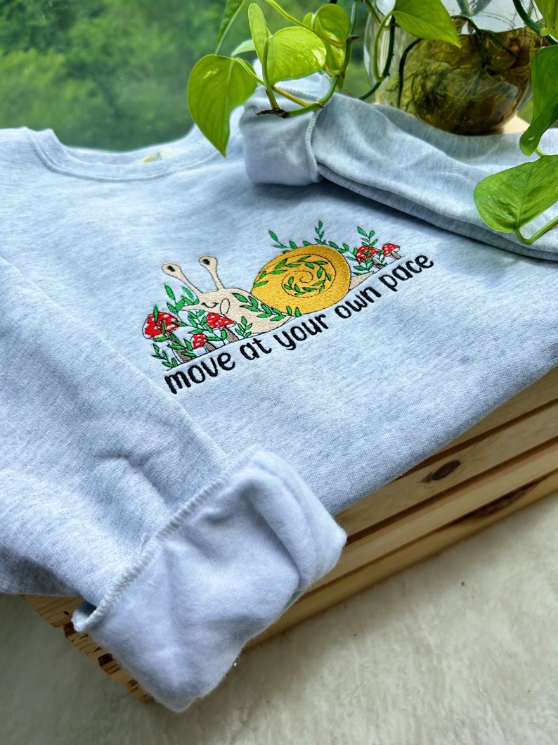 May include: A light gray sweatshirt with an embroidered snail design and the text "move at your own pace".
