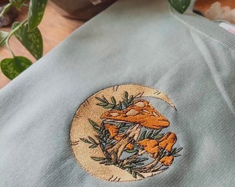 May include: A light blue sweatshirt with an embroidered design of mushrooms growing in a crescent moon. The mushrooms are orange and brown with white spots. The crescent moon is outlined in yellow.