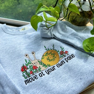 May include: A light blue sweatshirt with an embroidered snail design and the text "move at your own pace".