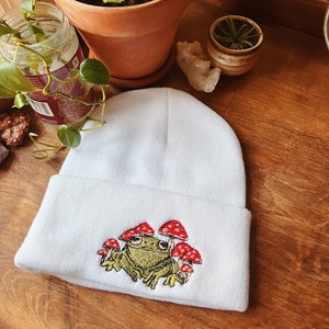 May include: White knit beanie with an embroidered green frog sitting amongst red and white mushrooms.