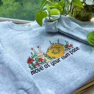 May include: A light blue sweatshirt with a snail and mushrooms embroidered on the front. The snail is brown and yellow and has a green shell. The mushrooms are red and white. The text "move at your own pace" is embroidered below the snail.