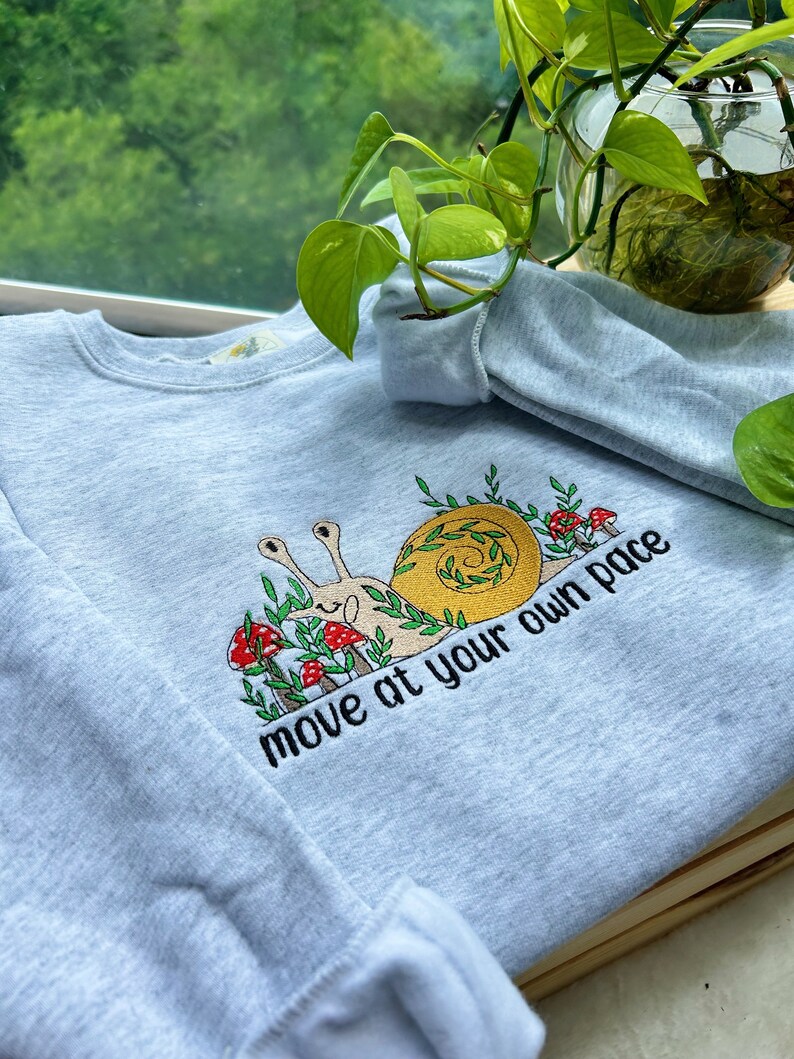 May include: A light blue sweatshirt with a snail and mushrooms embroidered on the front. The snail is brown and yellow and has a green shell. The mushrooms are red and white. The text "move at your own pace" is embroidered below the snail.
