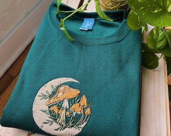 May include: A dark green sweatshirt with an embroidered design of mushrooms growing in a crescent moon. The design is in shades of yellow, brown, and green.