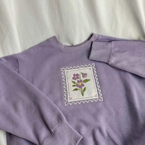 Peut inclure: Un sweat-shirt lavande avec un motif de timbre floral. Le timbre brodé présente des fleurs violettes avec des feuilles vertes sur un fond blanc, encadré par une bordure festonnée. Le sweat-shirt a des poignets côtelés et un col rond.