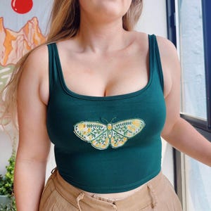 May include: A dark green vest top with a scoop neckline. The top has an embroidered butterfly design in yellow and white. The model is wearing tan trousers. The background includes a window and a painting.