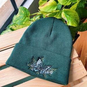 May include: A dark green beanie with an embroidered blue butterfly and floral design.