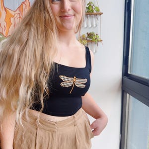 May include: Black crop top with a gingham dragonfly design. The top is paired with tan-colored pants. The image is taken indoors, with a window and decorative wall hanging in the background.