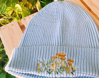 Puede incluir: Un gorro de punto azul claro con un diseño de bordado floral en amarillo y verde. El gorro está doblado por la mitad y se encuentra sobre una superficie de madera.