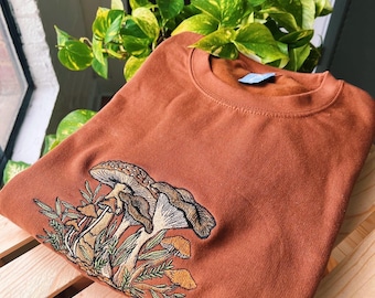 May include: A rust-colored sweatshirt with an embroidered design of mushrooms and foliage. The mushrooms are brown and white, and the foliage is green.