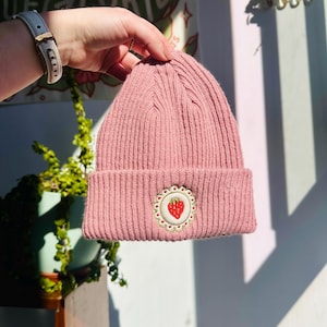 May include: A pink ribbed knit beanie with a decorative patch featuring a red strawberry. The beanie is held up in front of a white wall. The patch is round with a scalloped edge.