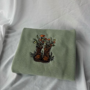 Puede incluir: Sudadera verde salvia con un diseño bordado de botas marrones llenas de flores naranjas y amarillas. Las botas están detalladas con costuras y las flores tienen hojas verdes. La sudadera está doblada sobre una superficie blanca.