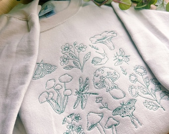 May include: A white sweatshirt with a green embroidered design of various nature elements, including mushrooms, flowers, butterflies, and dragonflies.