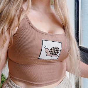 May include: A brown ribbed vest top with a square patch featuring a cartoon snail with a checkered shell. The top has a scoop neckline and is made of a soft, stretchy material.