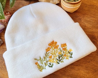 Puede incluir: Gorro blanco con flores amarillas y hojas verdes bordadas.