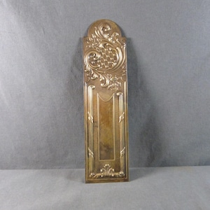 Op de afbeelding: Een rechthoekige, antieke, messingkleurige decoratieve plaat met sierlijke bloemen- en krulwerkontwerpen. De bovenkant heeft een afgeronde boog met gedetailleerde gravures, de onderkant een vergelijkbaar, kleiner ontwerp. Het midden heeft een verzonken paneel.