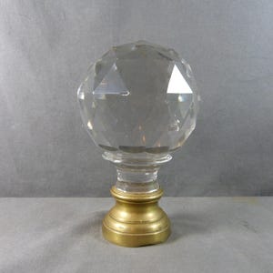 May include: A clear, multifaceted glass sphere finial mounted on a circular, brushed brass-toned metal base. The top section features a geometric, diamond-cut pattern that reflects light, while the base has a smooth, tiered, and rounded design.