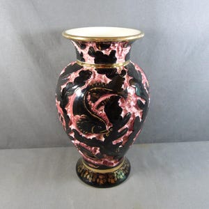 Könnte beinhalten: Eine dekorative Vase mit kontrastreichem Design, bestehend aus glänzend schwarzen Reliefmustern von Fischen und Korallen auf einem strukturierten, matten rosa Hintergrund. Der Rand und der Fuß sind goldfarben abgesetzt, mit Blumenmotiven am Sockel.
