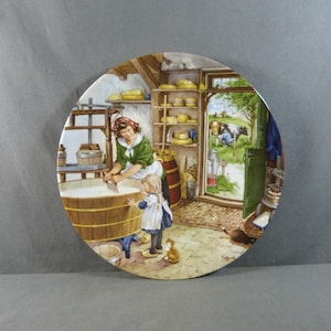 Op de afbeelding: Ronde decoratieve plaat met een gedetailleerde illustratie van een kaasbereidingsscène. Een vrouw en een kind worden afgebeeld in een rustiek gebouw, met planken met kaas en een uitzicht op een koe buiten.