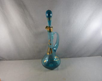 May include: A teal glass decanter with a handle and stopper. The decanter has gold leaf accents and a gold rim. The stopper is teal with gold stripes. The decanter is approximately 12 inches tall.