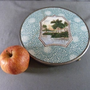Pode incluir: Uma bandeja redonda de metal com borda prateada e superfície azul-clara com motivos de pinhas brancas. Um painel hexagonal central exibe uma paisagem pintada de árvores perto de um corpo de água. Uma maçã vermelha está ao lado da bandeja.
