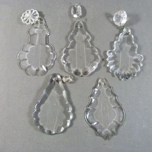 May include: Five clear glass chandelier crystals with scalloped edges and teardrop shapes. Three of the crystals feature small, round, faceted glass connectors at the top, while two have simple holes for hanging. The glass is transparent and smooth.