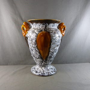 Peut inclure: Un vase décoratif en céramique avec une surface texturée et à motifs dans les tons de blanc, de gris et de marron. Le vase comporte deux poignées orange et un accent marron central. Le bord et l'intérieur sont également marron. Le vase a une base en piédestal.