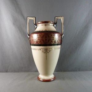 Peut inclure: Un vase crème avec une base évasée et deux poignées argentées. Le vase présente une bande rouge foncé avec des motifs floraux dorés et des accents noirs. Le bord et la base sont également rouge foncé.