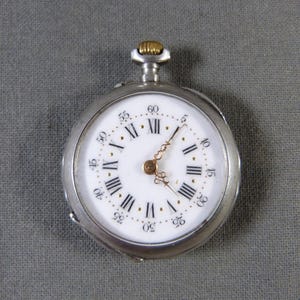 Peut inclure: Une montre de poche en argent avec un cadran blanc et des chiffres romains noirs. La montre a une tige de remontage et des aiguilles dorées.