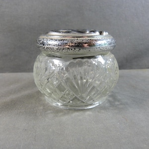 May include: A clear glass jar with a silver-colored lid. The jar has a round shape with a diamond-cut pattern. The lid is silver with a decorative design. The jar is likely for storing small items.