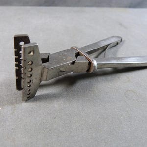 Könnte beinhalten: Ein vintage Werkzeug aus Metall mit einem T-förmigen Kopf, der entlang der Kante eine Reihe abgestufter kreisförmiger Löcher aufweist. Das Gerät hat zwei klappbare Griffe, die durch ein dünnes Metallband zusammengehalten werden.