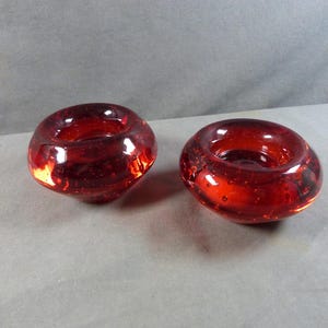 Op de afbeelding: Twee rode glazen kandelaars. De kandelaars zijn rond met een brede opening en een enigszins onregelmatige vorm. Het glas heeft een bubbelachtige textuur en een dieprode kleur, met enkele lichtere plekken.