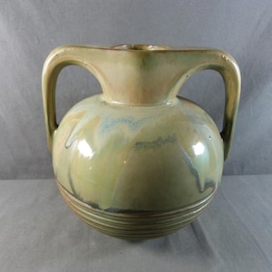 Pode incluir: Um vaso de cerâmica com corpo arredondado e duas alças arqueadas. O vaso tem um acabamento brilhante com tons de verde, bege e azul. Linhas horizontais circundam a base. Esta peça decorativa é adequada para decoração de casa.