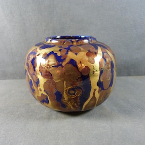 Könnte beinhalten: Eine runde Keramikvase mit einem gold-, blau- und braun melierten Design. Die Vase hat einen breiten Körper und eine schmale Öffnung mit blauem Rand. Die Goldbereiche haben einen metallischen Glanz.