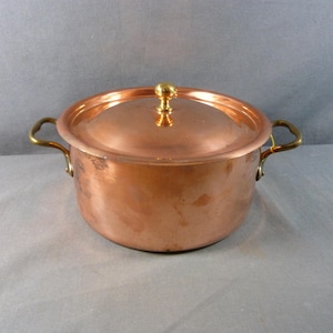 May include: A copper cooking pot with a lid and brass handles. The pot has a round shape and a shiny, reflective surface. The lid is topped with a brass knob. The pot is designed for cooking and food preparation.