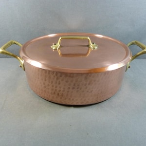 May include: A copper pot with a lid and brass handles. The pot has a hammered texture on the sides and a brass handle on the lid. The pot is round and sits on a gray surface.