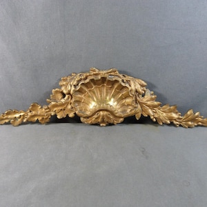 Könnte beinhalten: Ein goldfarbenes Wandornament mit einem zentralen, muschelförmigen Motiv, das von symmetrischen, länglichen Akanthusblatt-Schnitzereien flankiert wird. Das Dekorationsobjekt hat eine verwitterte, metallische Oberfläche mit detaillierten Reliefs.