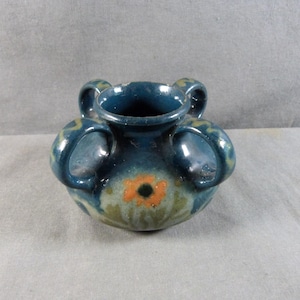 May include: A small, round, ceramic vase with a dark teal glaze. The vase has two handles and a floral design in orange and green. The vase is approximately 10 cm wide and 8 cm tall.